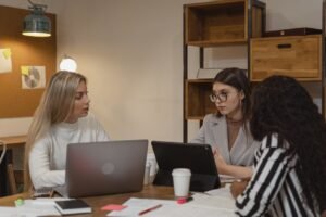 Three women engaging in a focused discussion in an office setting with laptops and documents.